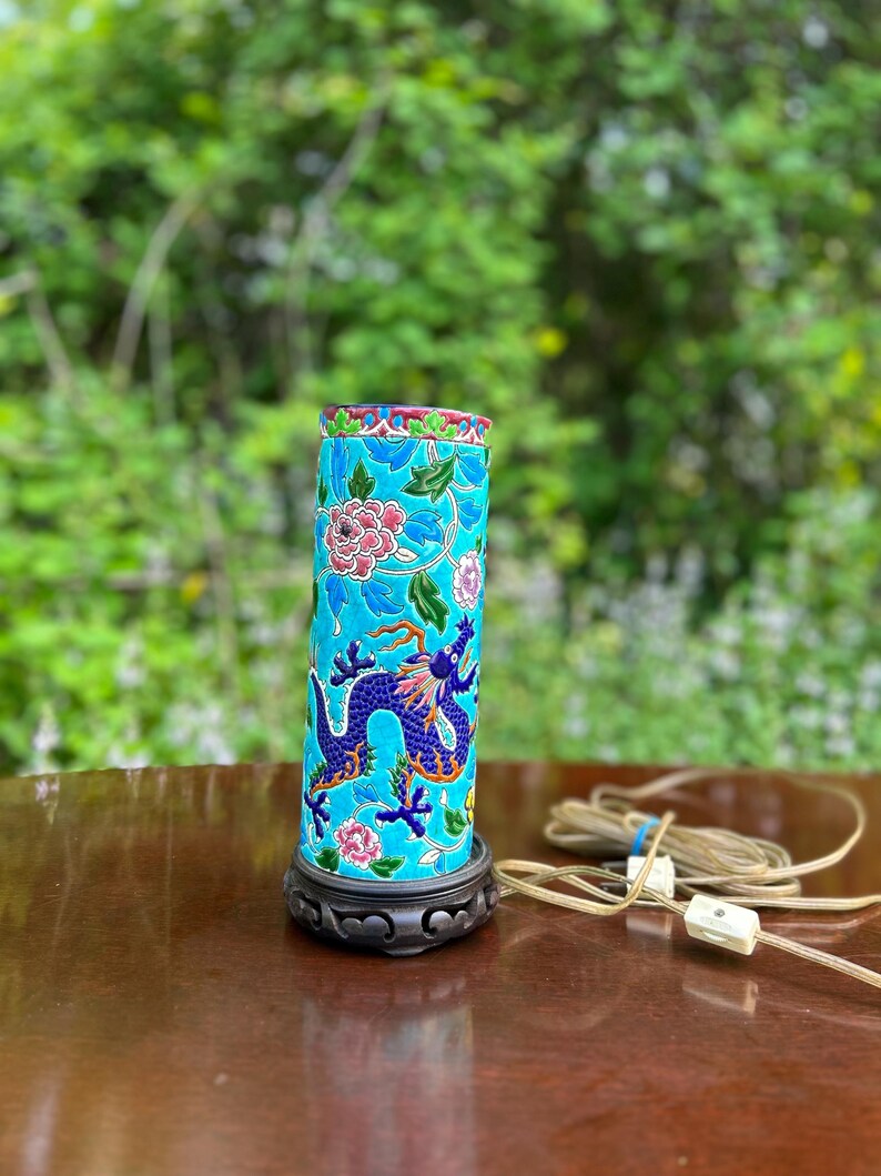 Puede incluir: Una l&aacute;mpara de mesa de cer&aacute;mica azul y verde con un dise&ntilde;o de drag&oacute;n y flores. La l&aacute;mpara tiene una base de madera y un cable con un interruptor blanco.
