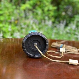 Puede incluir: La parte inferior de una base de l&aacute;mpara negra con un cable y un enchufe. La base tiene un dise&ntilde;o circular texturizado.