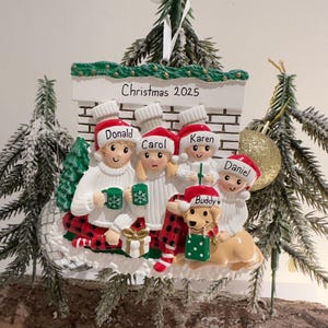 May include: A Christmas ornament featuring a family of five and a dog, all wearing Santa hats and holding mugs. The ornament has a fireplace design with the text "Christmas 2025". The family is dressed in white sweaters and red plaid accents.