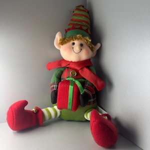 Pode incluir: Uma boneca de duende de Natal de pelúcia com um rosto sorridente, vestindo uma jaqueta xadrez verde e vermelha, um cachecol vermelho e um chapéu listrado verde e branco. O duende segura uma caixa de presente vermelha com uma fita verde e tem sapatos vermelhos com um pequeno sino dourado.
