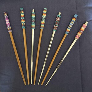 May include: A collection of chopsticks with beaded tops in various colors and patterns. Some chopsticks are bamboo, while others are metal. The beaded sections feature designs in shades of blue, orange, yellow, and pink.