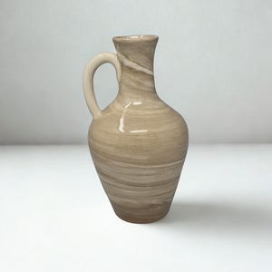 May include: A beige ceramic pitcher with a handle. The pitcher has a rounded body and a narrow neck. The surface has a swirled pattern of light and dark beige tones. The handle is a solid, lighter beige color.