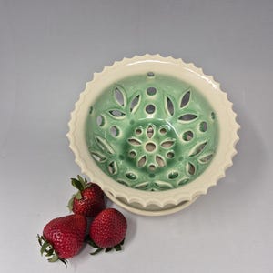 May include: A ceramic fruit bowl with a green interior and a cream-coloured rim. The bowl has a decorative floral pattern with cut-out designs. Three fresh red strawberries are next to the bowl.