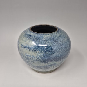 Puede incluir: Un pequeño jarrón redondo de cerámica con un esmalte moteado azul y blanco. El jarrón tiene una abertura ancha y un fondo redondeado.