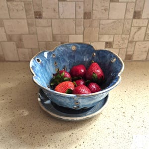 May include: A blue ceramic fruit bowl with a scalloped edge, filled with fresh red strawberries. The bowl sits on a matching saucer. The bowl has small holes around the rim. The background features a neutral-toned tile backsplash.