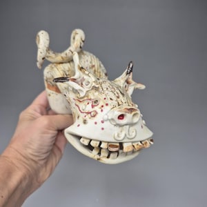 Könnte beinhalten: Eine skurrile Keramikskulptur, die ein Wesen mit breitem Grinsen und Zähnen darstellt. Die Skulptur ist überwiegend cremefarben mit braunen und roten Details, einschließlich Punkten und Gesichtsmerkmalen. Das Stück hat eine unverwechselbare, handgefertigte Ästhetik.