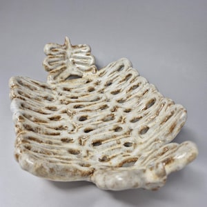May include: A handcrafted, cream-coloured ceramic dish with a distinctive openwork design. The dish has a textured surface and a rib-like pattern with small holes. The edges are slightly uneven, and it features a small decorative element at the top.