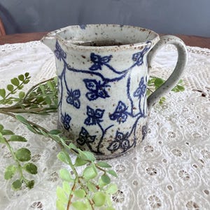 May include: A set of three ceramic mugs with a speckled, cream-colored glaze. Each mug features a different hand-painted floral design in shades of blue, green, and yellow. The mugs have a simple, rounded shape and a comfortable handle.