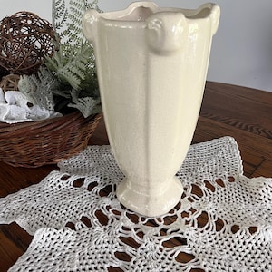 May include: A white ceramic vase with a fluted design and a scalloped rim. The vase has a textured surface and a slightly flared base.
