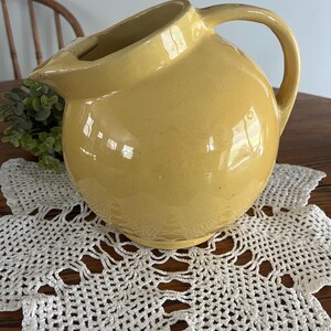 May include: A yellow ceramic pitcher with a rounded body and a handle. The pitcher has a spout on the side and a smooth, glossy finish.
