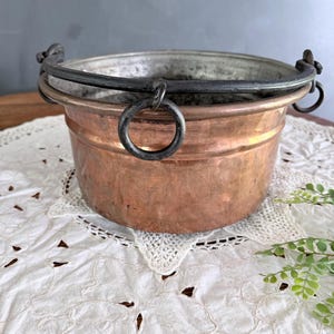 May include: A round copper pot with a hammered texture and a wide, flared rim. It features a dark, heavy iron swing handle attached to two side rings. The interior of the pot has a metallic, silver-toned lining, showing signs of wear and age.