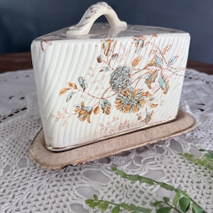 May include: A triangular ceramic butter dish with a cream-colored base and a matching lid featuring a diagonal ribbed texture. The lid is decorated with a floral pattern in shades of blue, orange, and brown, and includes a curved handle on top.