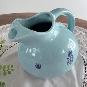 May include: A light blue ceramic pitcher with a rounded body and a curved handle. The pitcher has a spout and is decorated with two dark blue floral designs. The pitcher is sitting on a white lace doily.