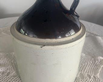 May include: A vintage stoneware jug with a dark brown glazed top and handle, contrasting with a white base. The jug has a rounded body and a small spout. The handle is arched, and the jug appears to be in good condition.