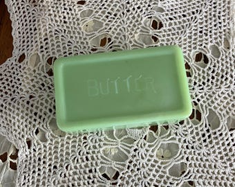 May include: A rectangular, light green butter dish with the word "BUTTER" etched on top. The dish sits on a white, crocheted doily with a floral pattern. The background is a dark wooden surface.