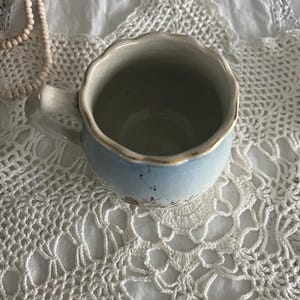 Pu&ograve; includere: Una tazza in ceramica blu chiaro con un bordo ondulato e un motivo floreale. La tazza &egrave; appoggiata su un centrino bianco all'uncinetto.