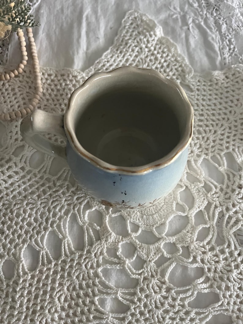 Pu&ograve; includere: Una tazza in ceramica blu chiaro con un bordo ondulato e un motivo floreale. La tazza &egrave; appoggiata su un centrino bianco all'uncinetto.