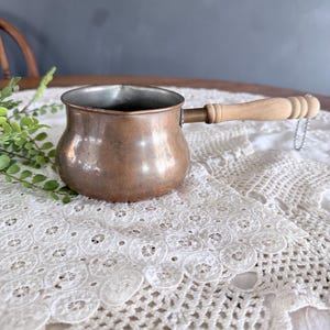 May include: A copper-colored Turkish coffee pot with a wooden handle and a small chain. The pot has a rounded body and a spout. The handle is attached with a brass fitting. The pot is sitting on a white lace tablecloth.