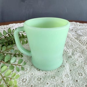 May include: A vintage jadeite glass mug with a handle. The mug has a smooth, slightly speckled surface and a light green color. It is set on a white lace tablecloth with a sprig of green leaves.