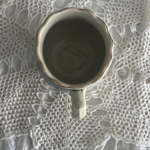 Pu&ograve; includere: Una tazza in ceramica bianca con un bordo ondulato e un bordo dorato. La tazza ha un manico ed &egrave; appoggiata su una superficie di uncinetto bianca.