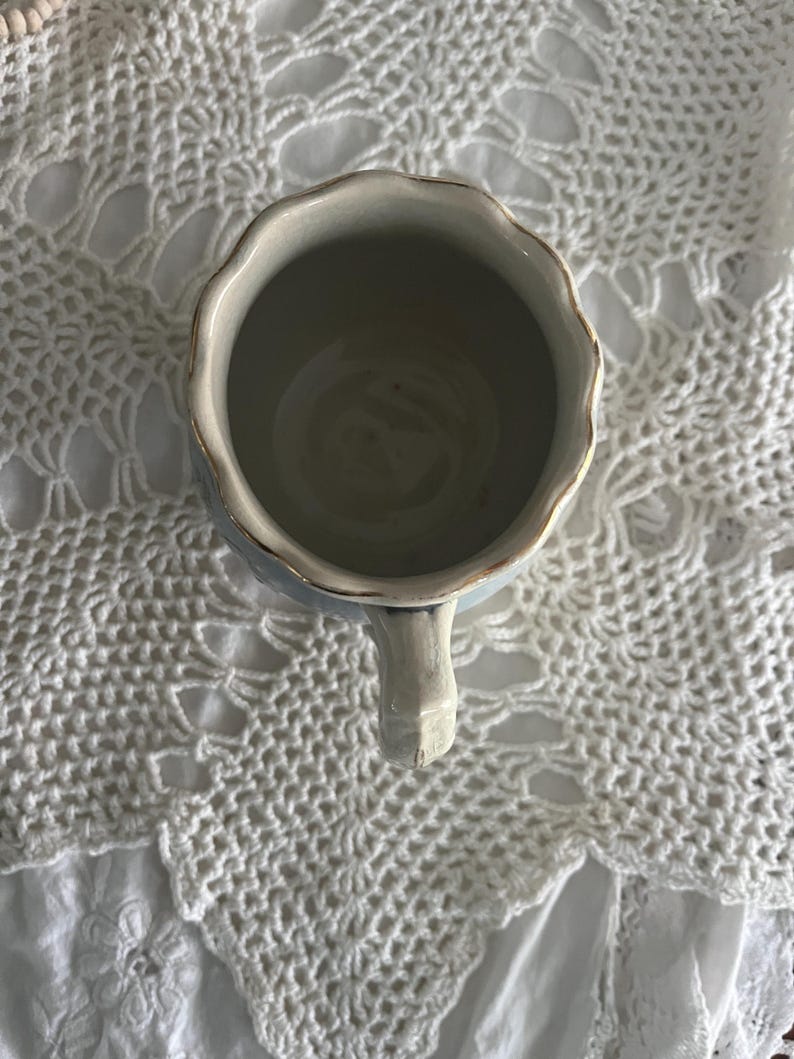 Pu&ograve; includere: Una tazza in ceramica bianca con un bordo ondulato e un bordo dorato. La tazza ha un manico ed &egrave; appoggiata su una superficie di uncinetto bianca.