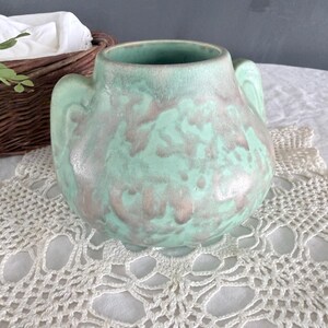 May include: A ceramic vase with a rounded body and two small handles. The vase has a mottled finish in shades of seafoam green and gray. It sits on a white crocheted doily.