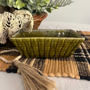 May include: A small, rectangular, green ceramic dish with a woven pattern. The dish is sitting on a woven rug with black and white stripes.