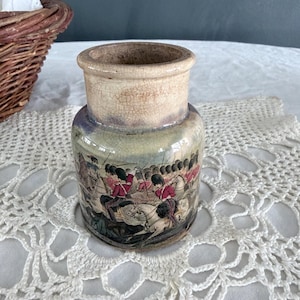 May include: A small, antique ceramic jar with a detailed illustration of soldiers in red coats on horseback. The jar has a beige, cracked finish and a wide, unglazed rim. The base color is a pale green.