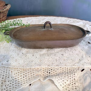 May include: An antique oval-shaped metal casserole dish with a lid. The dish is a warm bronze color with a matching handle on the lid. The dish has handles on each side. The dish is sitting on a white lace tablecloth.