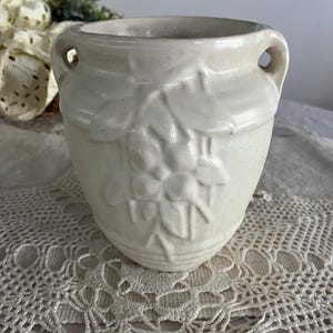May include: Off-white ceramic vase with two small handles and a floral relief design. The vase has a slightly textured surface and a rounded shape. The background includes a lace doily and some greenery.