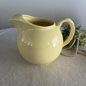 May include: A pale yellow ceramic pitcher with a rounded body and a curved handle. The pitcher is sitting on a white crocheted placemat.