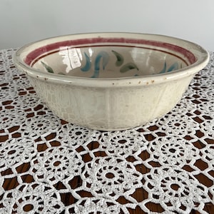 May include: Cream-colored ceramic bowl with a wide rim and a decorative painted design. The rim features a maroon stripe, and the interior has blue and green abstract shapes. The bowl is sitting on a white lace doily.