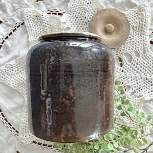 May include: A cylindrical dark brown ceramic storage jar with a glossy, mottled glaze finish. The jar features a wide opening and a separate, circular, light-colored, unglazed ceramic lid with a small central knob handle for easy removal.
