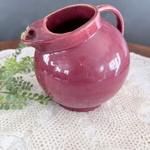 May include: A round, glossy, burgundy-colored pitcher with a handle and spout. The pitcher is sitting on a white lace doily. The pitcher is a vintage piece.