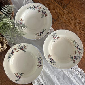 May include: Three white ceramic soup bowls with blue and red floral and bird designs. The bowls have a scalloped edge and are on a white lace tablecloth.