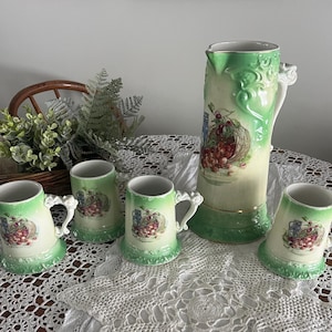 May include: A set of five vintage ceramic mugs and a pitcher with a green and white floral design featuring cherries. The mugs and pitcher have a green glaze and gold accents.