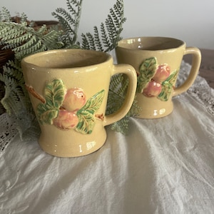 May include: Two vintage ceramic mugs with a peach and leaf design. The mugs are a pale yellow color with green leaves and pink peaches.