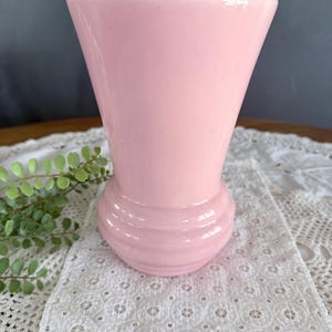 May include: A light pink vase with a flared top and a rounded base. The vase has a smooth, glossy finish and is approximately 7 inches tall. The base features horizontal ridges. The vase is sitting on a white lace tablecloth.