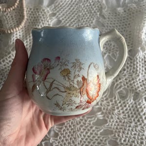 Pu&ograve; includere: Un'antica brocca per la panna in porcellana con una glassa blu e bianca. La brocca presenta un motivo floreale con fiori arancioni e rosa e foglie verdi.