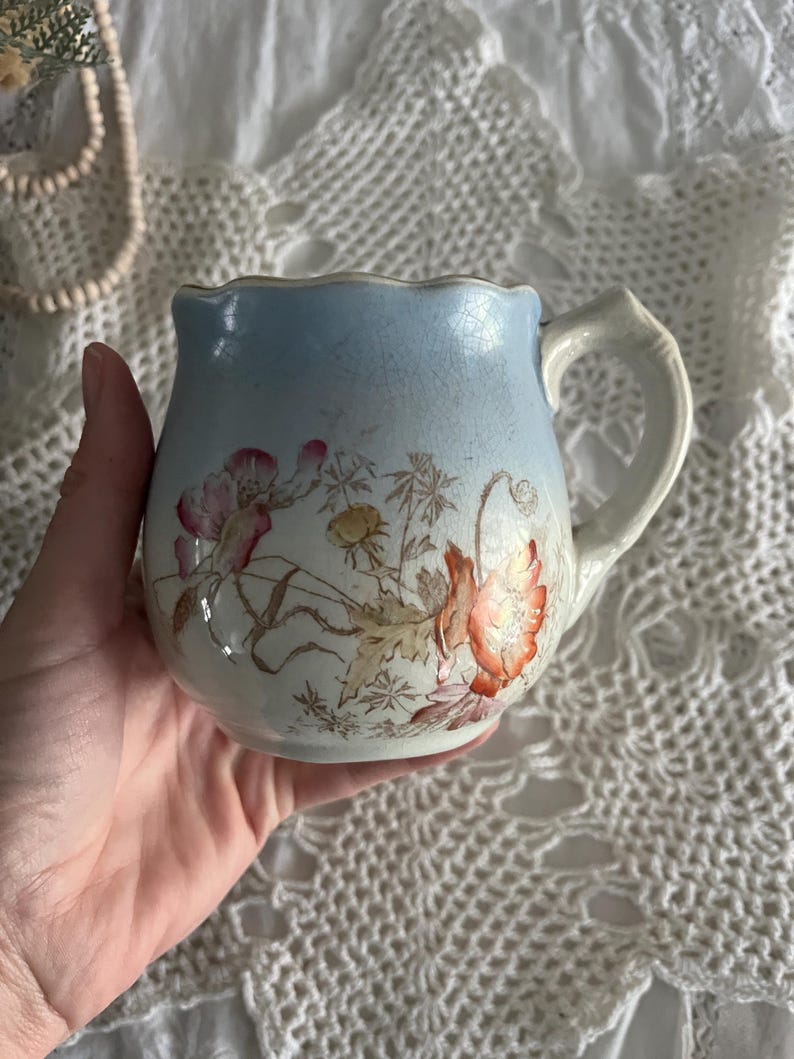 Pu&ograve; includere: Un'antica brocca per la panna in porcellana con una glassa blu e bianca. La brocca presenta un motivo floreale con fiori arancioni e rosa e foglie verdi.