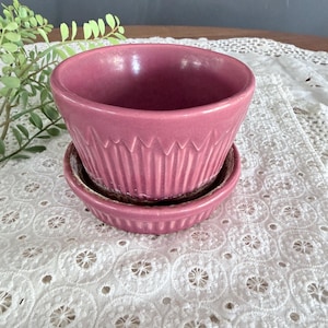 May include: A small, round, pink ceramic planter with a matching saucer. The planter has a ridged design around the exterior. The saucer is a smaller, shallow dish. The set is likely for small plants or succulents.