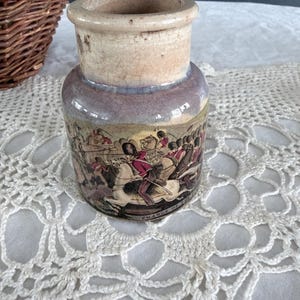 May include: A small, antique ceramic jar with a wide opening and a faded, multi-toned exterior. The jar features a detailed illustration of a historical battle scene around its base. The text "CHARGE OF THE" is visible on the jar.