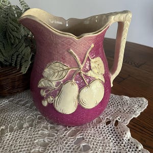 May include: A vintage pink and white ceramic pitcher with a pear design. The pitcher has a handle and a wide mouth.