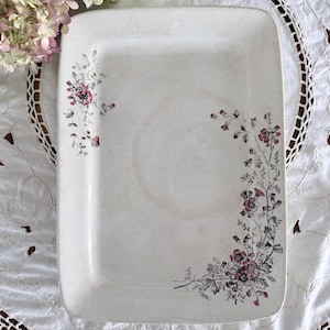 May include: A rectangular, off-white ceramic serving platter with delicate floral designs in pink and gray. The flowers are clustered in the corners of the platter. The platter is placed on a white lace tablecloth.