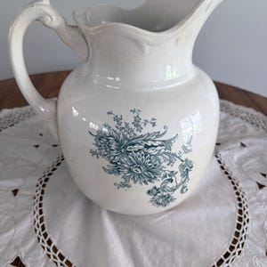 May include: White ceramic pitcher with a floral design in teal. The pitcher has a curved handle and a spout. The pitcher is sitting on a white doily with a decorative edge. The pitcher is a vintage style.