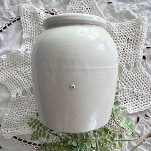 May include: A cylindrical off-white ceramic crock featuring a smooth, glazed finish with a visible network of fine surface crazing. The vessel has a thick, rounded rim at the top and a small, singular chip located on the center of the body.