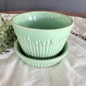 May include: A light green ceramic planter and matching saucer. The planter has a ridged design around the sides and a smooth interior. The saucer has a similar ridged design. The set is made of ceramic.