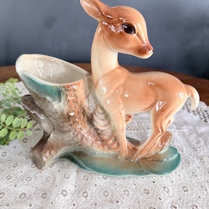 May include: A ceramic deer planter in shades of brown, tan, and cream. The deer stands next to a tree stump with a planter opening. The base is a teal color. The deer has dark brown eyes and a glossy finish.