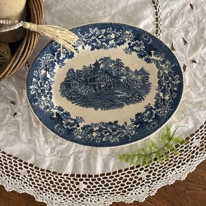 May include: A blue and white ceramic plate with a floral border and a scene of a house in a landscape. The plate is on a white crocheted tablecloth.