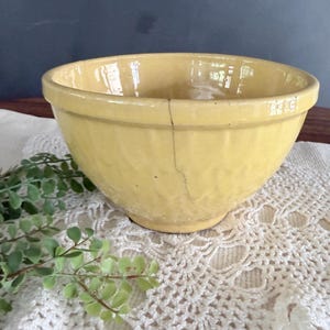 May include: A yellow ceramic mixing bowl with a glossy finish and a textured exterior pattern. The bowl features a prominent vertical crack extending from the top rim down toward the base, showing signs of age and wear on its surface.